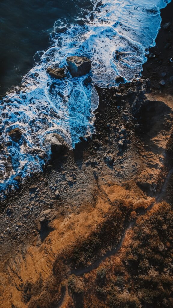 Aerial drone photograph by Wandering Ducks Photo & Film of rocky coast with aqua and indigo waves and rust and mauve beach and grasses